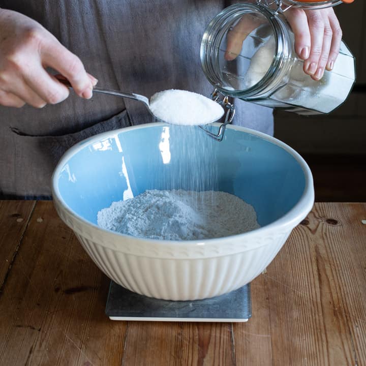 womans hands holding a metal spoon of sugar over a large blue and white mixing bowl