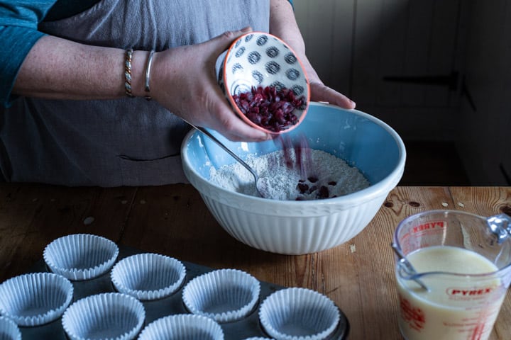 womans hands tipping a small bowl of cranberries into a large blue and white bowl of muffin mixture