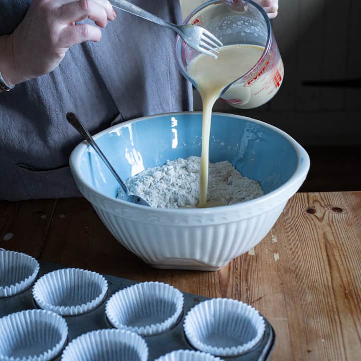 womans hands pouring wet ingredients into a bowl of flour next to a tin of muffin paper cases
