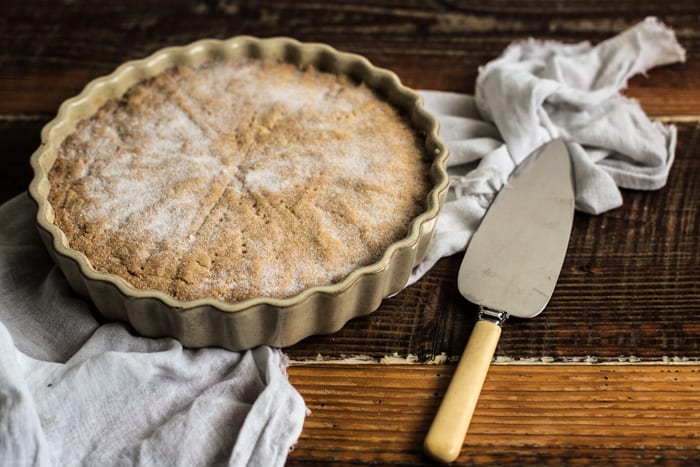 Wooden background with white plate and cake slice, and a whole cardamom shortbread dusted with sugar