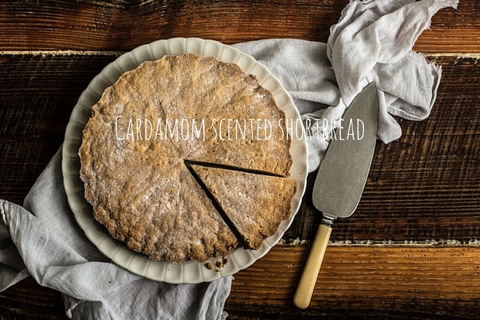 Wooden background with white plate and cake slice, and a whole cardamom shortbread dusted with sugar