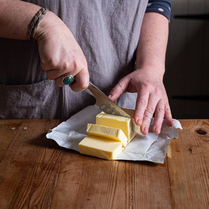 womnas hands cutting a block of butter on a wooden kitchen counter