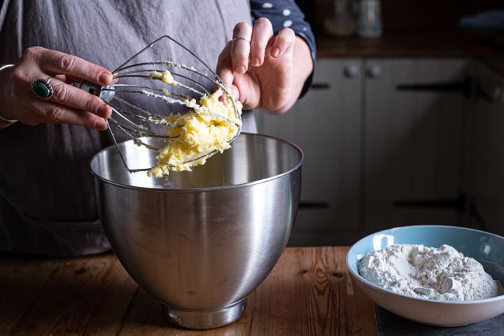womnas hands removing whipped sugar and butter from a whisk over a large silver mixing bowl 