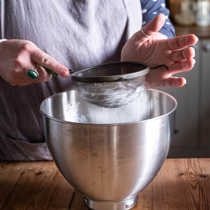 womans hands sifting flour into a large silver mixing bowl