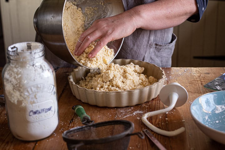 womans hands tipping shortbread mix into a flan dish on a wooden kitchen counter surrounded by baking mess