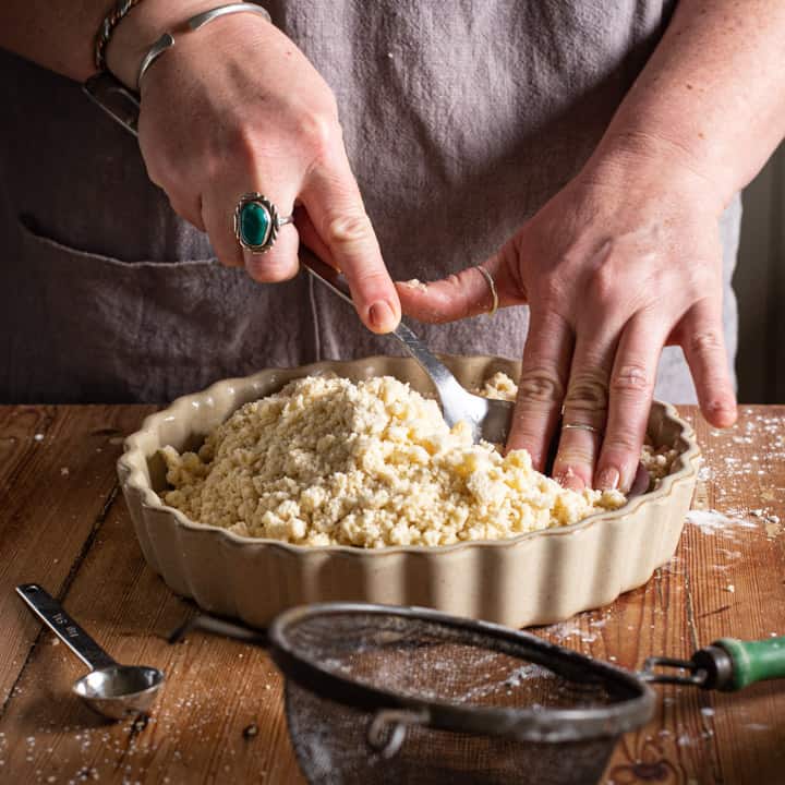 womans hands pressing shortbread mix into a flan dish on a wooden kitchen counter surrounded by baking mess