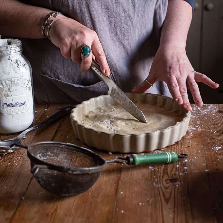 womans hands scoring shortbread in a flan dish on a wooden kitchen counter surrounded by baking mess