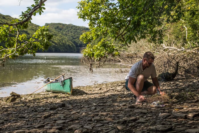 Man making fire on river bank with green canoe morrend behind him