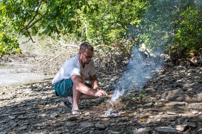 Man making a small campfire on the river bank with oak trees overhead