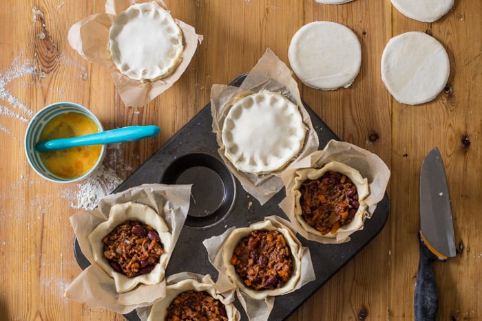 Chilli Beef Pies being assembled in a muffin tin on a wooden table surrounded by baking equipment