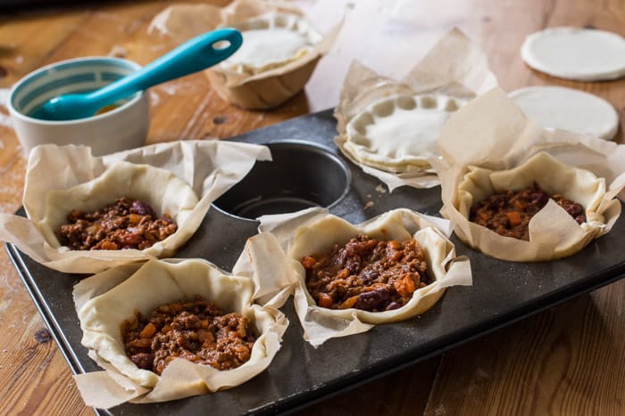 Chilli Beef Pies being assembled in a muffin tin on a wooden table surrounded by baking equipment