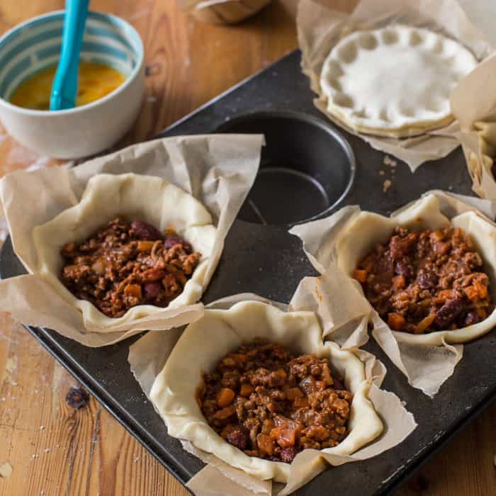 Chilli Beef Pies being assembled in a muffin tin on a wooden table surrounded by baking equipment