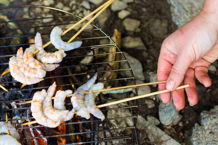 Woman’s hands placing king prawn kebabs onto a campfire grill