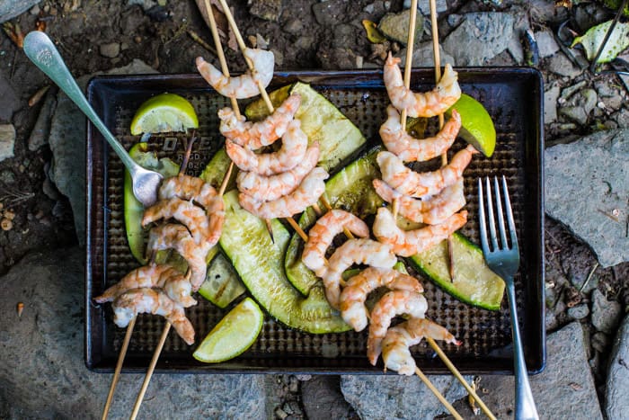 4 wooden skewers filled with pink juicy prawns on a baking tray with courgette ribbons and wedges of lime