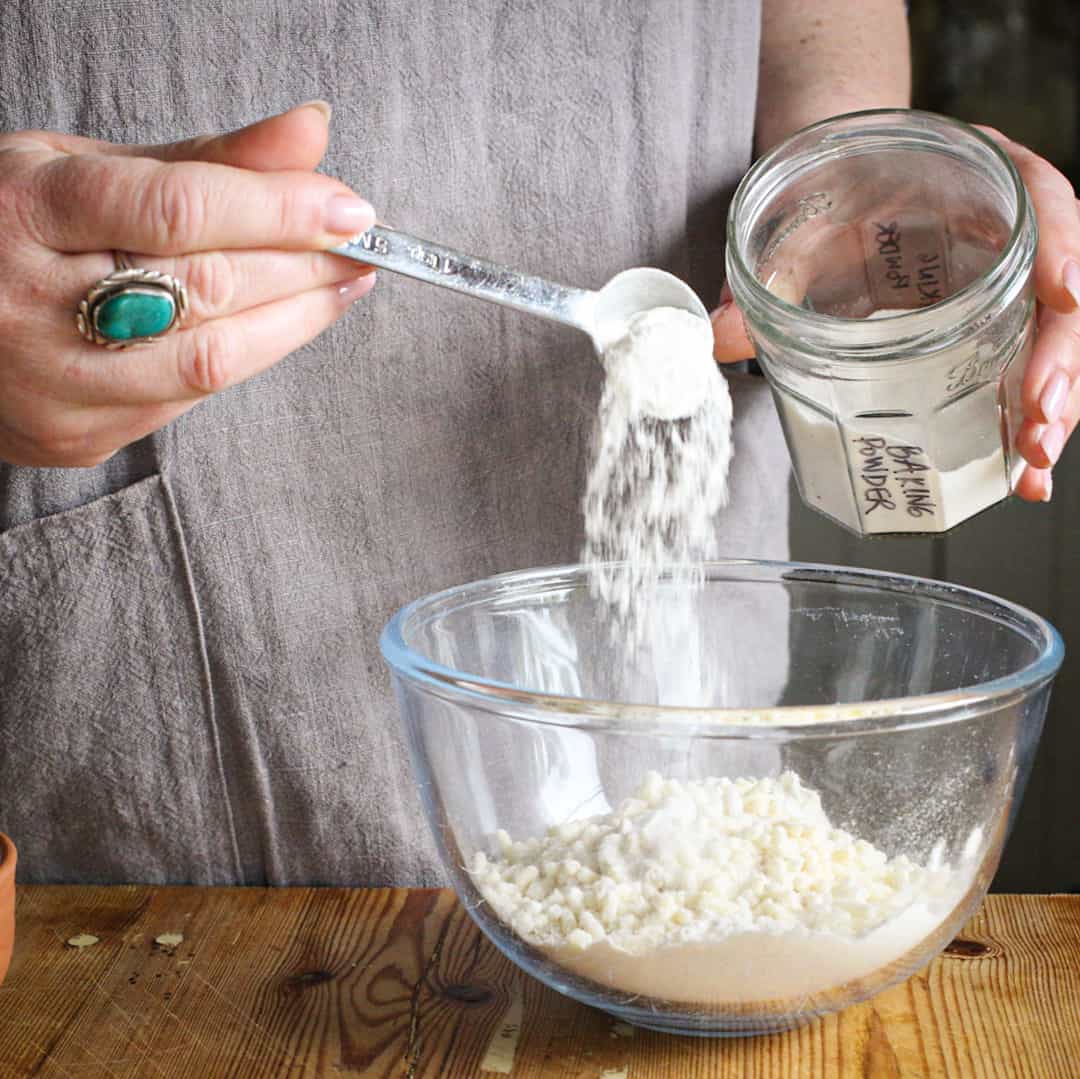 Woman’s hands spooning white baking powder into a glass bowl of flour