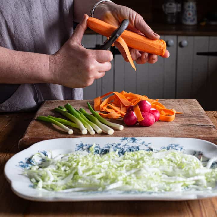 woman in grey grating a carrot over a bunch of other salad vegetables