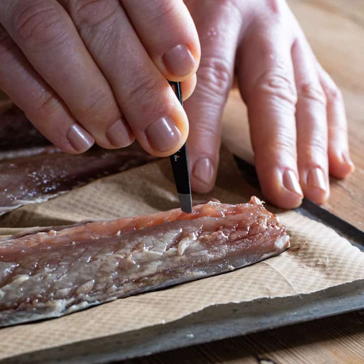 womans hands holding a pair of tweezers and pinching bones out of a fresh mackerel fillet