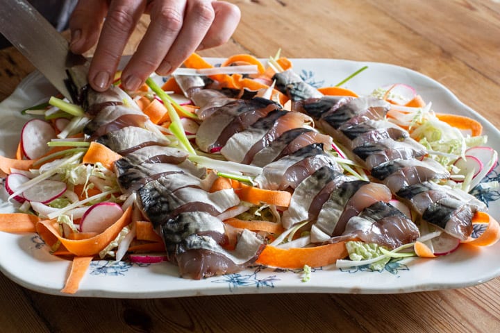 womans hands carefully placing slices of raw mackerel on top of a fresh salad on a blue and white plate