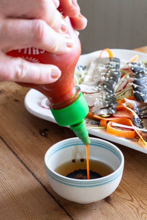 Sriracha hot sauce being poured into a small blue and white bowl next to a white plate mackerel sashimi and salad