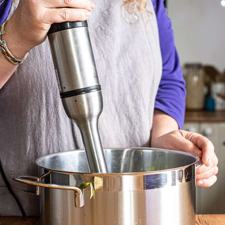 womans hands holding a stick blender in a stock pot of broccoli and almond soup