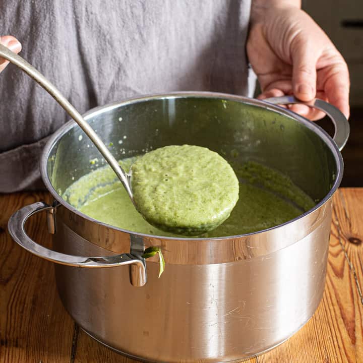 womans hands holding a ladle of bright green broccoli and almond soup