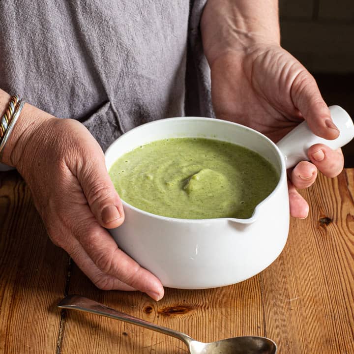 womans hands holding a white bowl of bright green broccoli soup against a wooden background