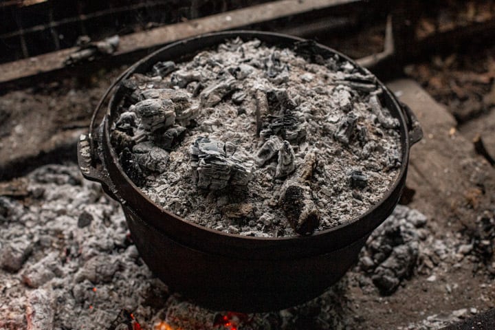 dutch oven sitting in campfire embers with more embers on the lid roasting a whole chicken