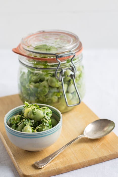  A kilner jar of Marinated Broad Beans beside a white bowl of beans and a spoon on a wooden board against a white background