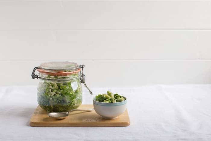  A kilner jar of Marinated Broad Beans beside a white bowl of beans and a spoon on a wooden board against a white background