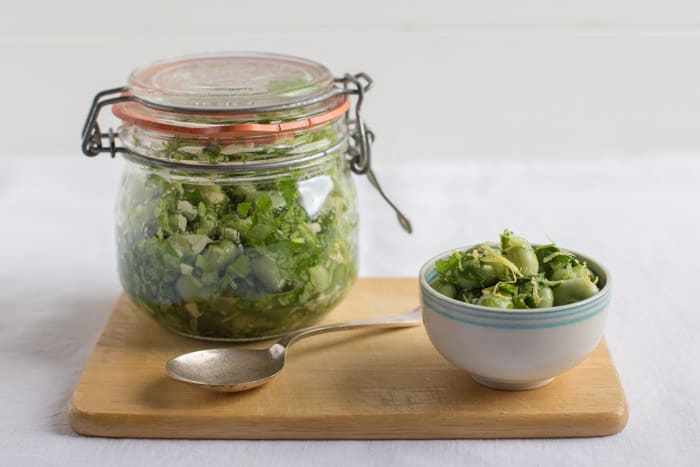  A kilner jar of Marinated Broad Beans beside a white bowl of beans and a spoon on a wooden board against a white background