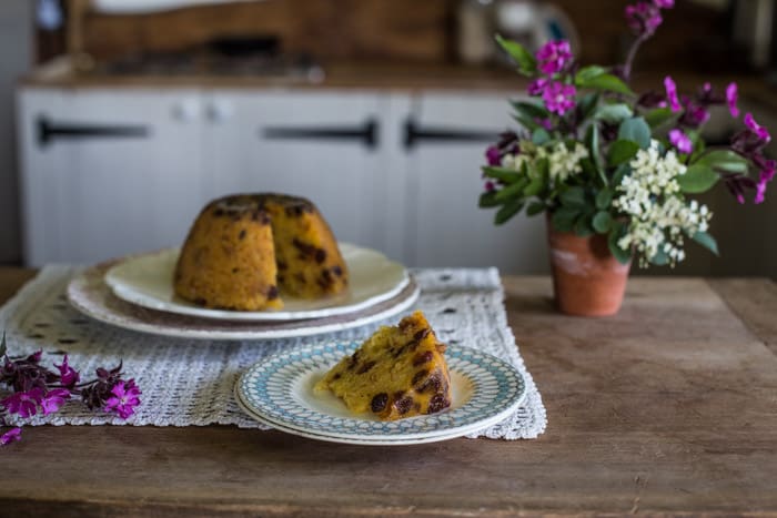 A slice of Snowdon Pudding on a white plate beside the rest of thepudding on a serving plate with a plant beside them