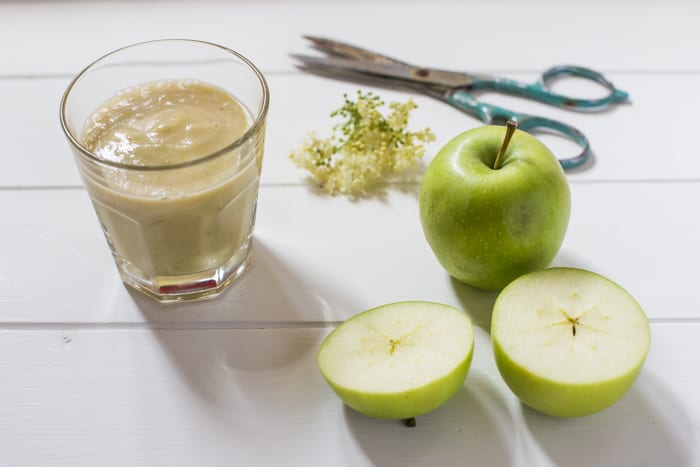 Apple and Elderflower Smoothie in a glass on a white table with apples , Elderflowers & scissors