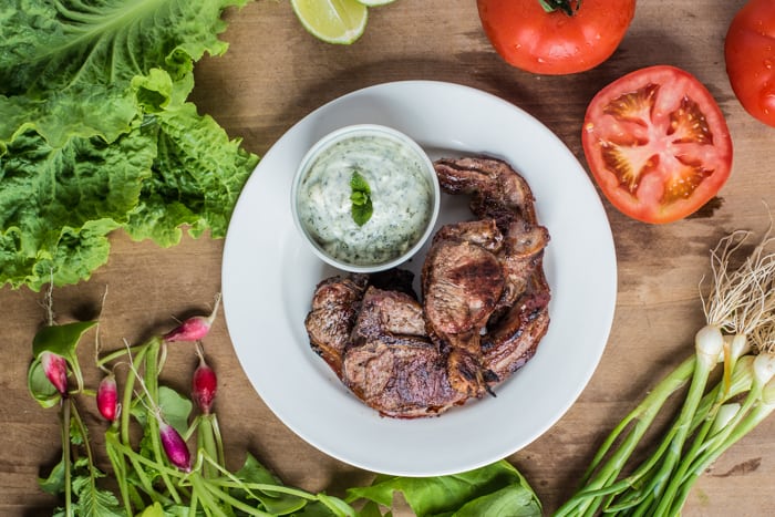 BBQ Lamb Chops with Minted Mayo on a white plate surrounded by salad ingredients on a wooden table