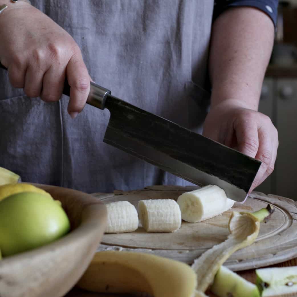 Woman in grey chopping a banana into a pieces on a wooden chopping board with a large knife