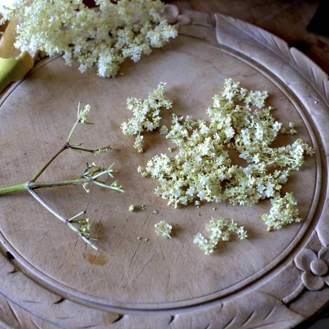 Wooden chopping board with an elderflower head that has had the tiny white blossom removed and piled up on the side