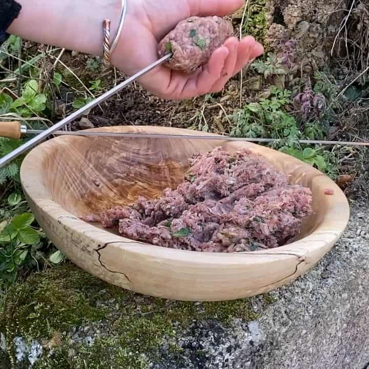 womans hands shaping beef kebabs onto a metal skewer from a wooden bowl next to a campfire