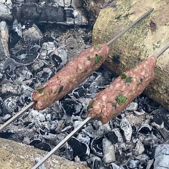 2 metal skewers with kebabs on cooking over the embers of a campfire