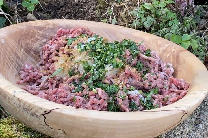 a wooden bowl with minced beef, herbs and spices outdoors next to a campfire ready to make kabobs