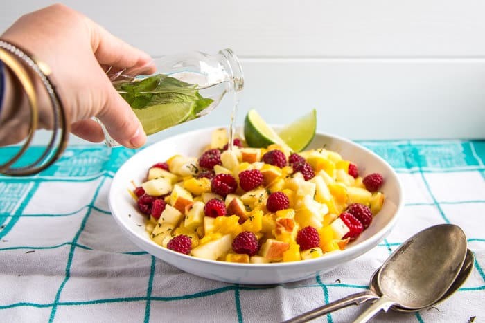 Mojito Fruit Salad Dressing being poured onto a white bowl of fruitbeside 2 spoons on a green and white checked cloth