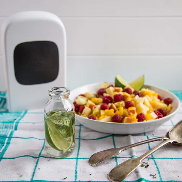 Mojito Fruit Salad Dressing in a glass bottle beside the vegetable chopper and a bowl of fruit and 2 spoons on a green and white checked cloth