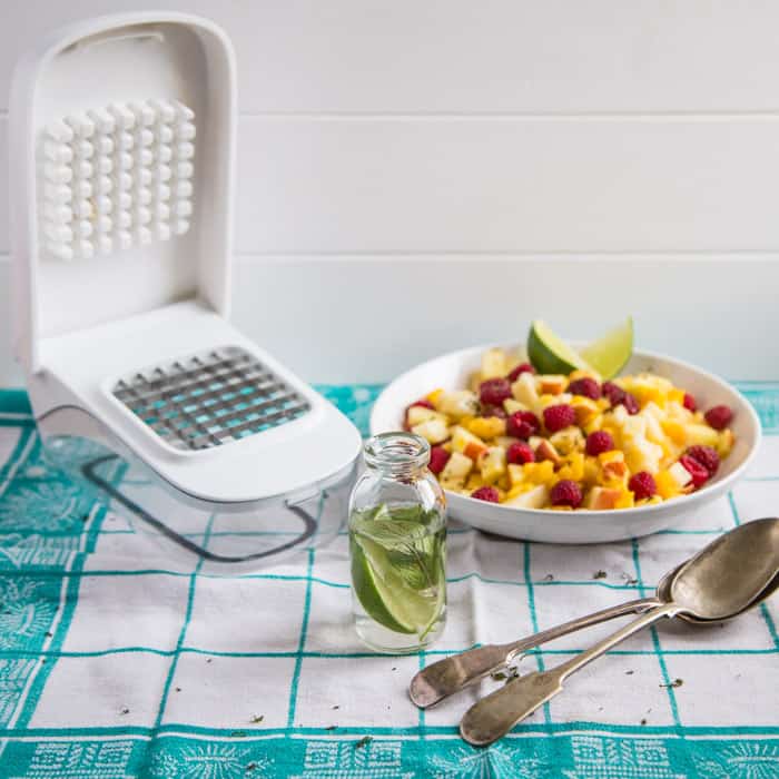 Mojito Fruit Salad Dressing in a glass bottle beside the vegetable chopper and a bowl of fruit and 2 spoons on a green and white checked cloth