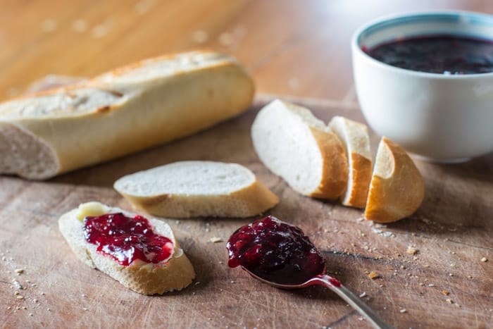 Sloe and wild blackberry hedgerow jam recipe spread on crusty bread surrounded by more bread a bowl of jam and a spoon on a wooden table