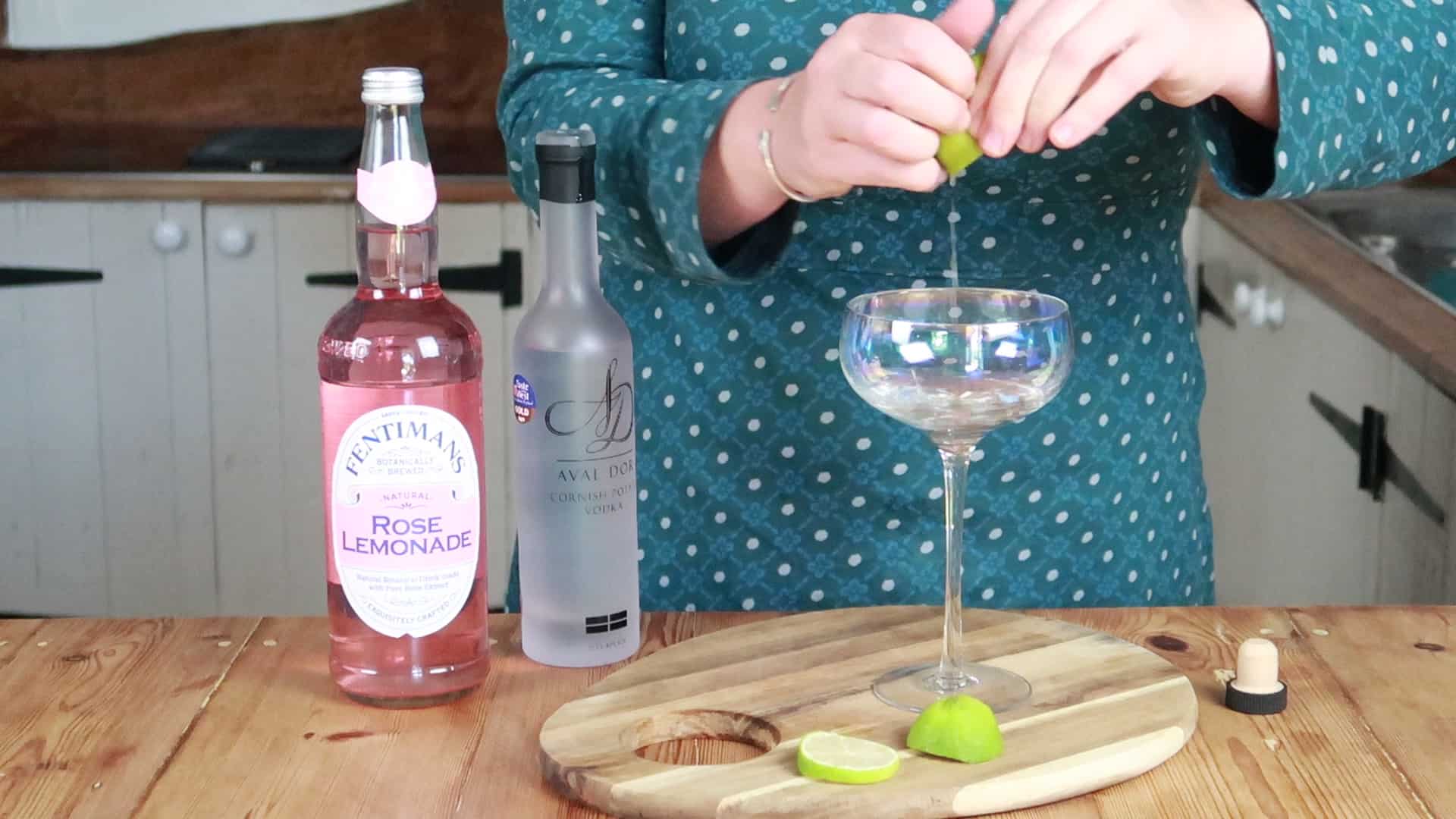 Person squeezing lime into a cocktail glass stood on a wooden board with bottle of lemonade and vodka beside it