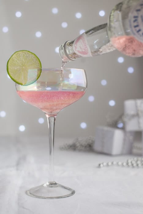 Pouring a glass of Pink Elephant Cocktail garnished with a slice of Lime and festive parcels on a grey and white background