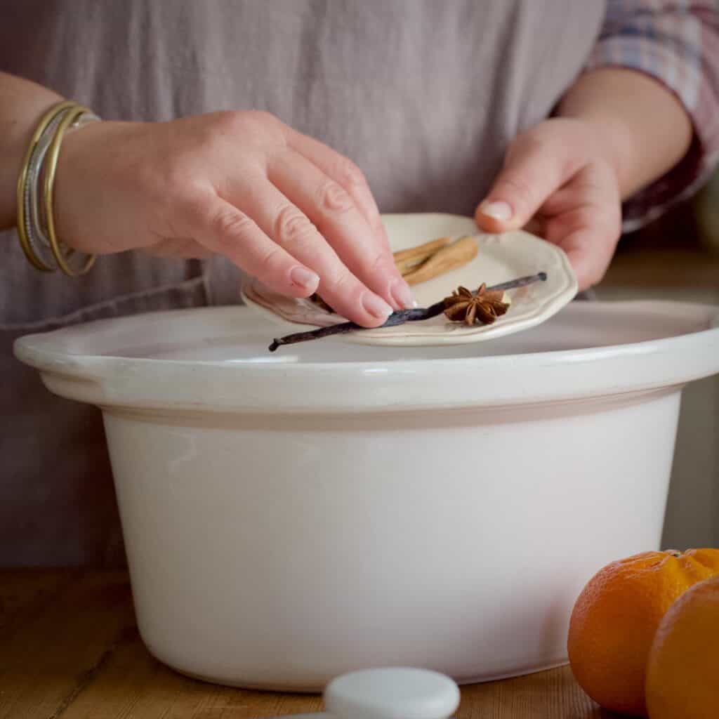 Womans hands moving spices from a small white plate into a large white slow cooker pot