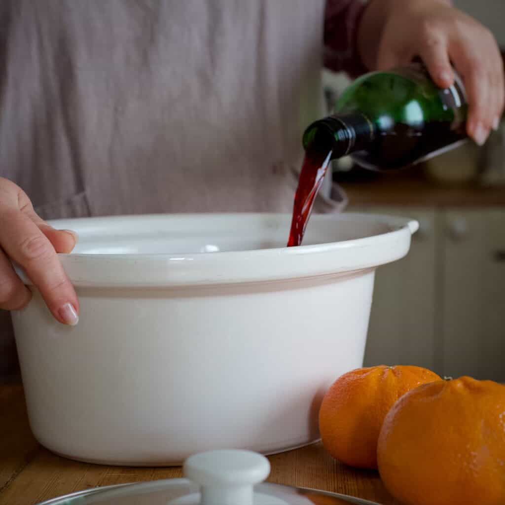 Woman in grey pouring red wine from a green glass wine bottle into a white slow cooker bowl