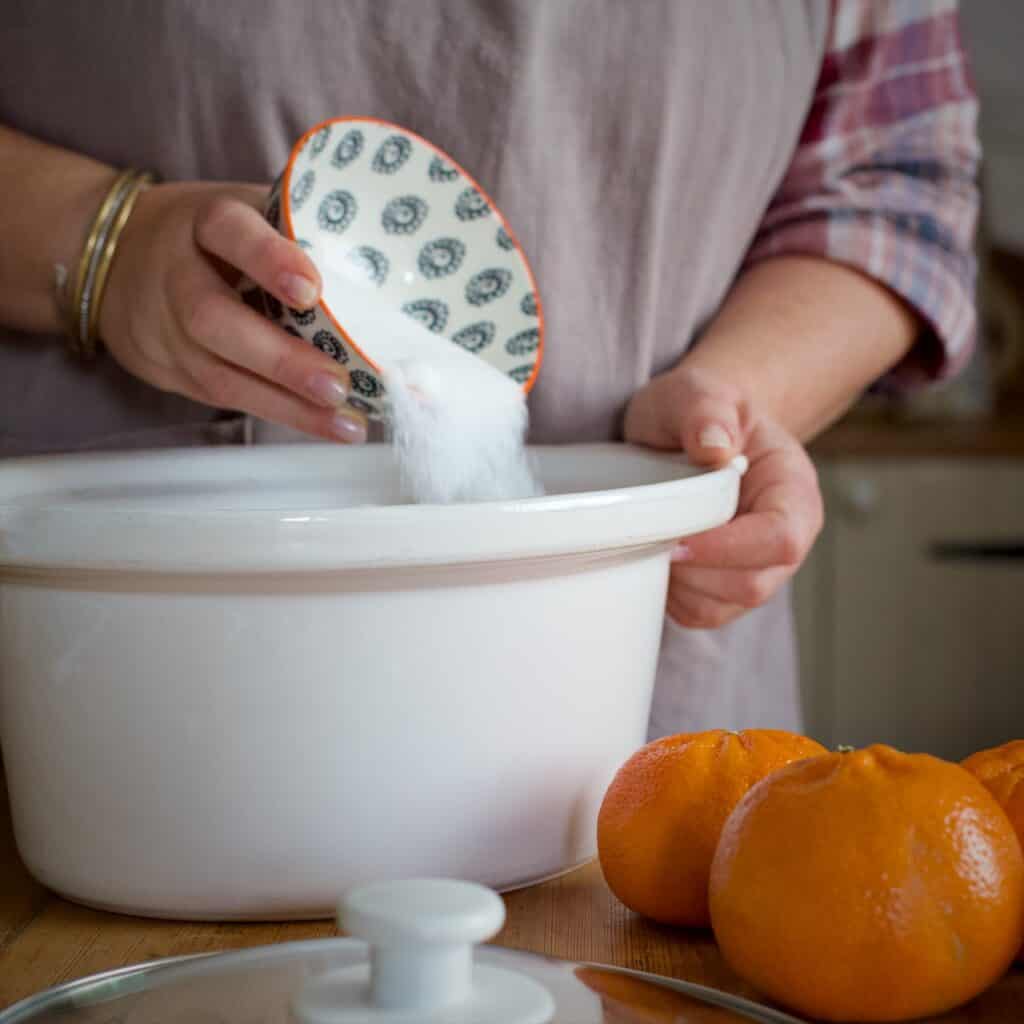 Womans hands pouring white sugar from a small black and white bowl into a white slow cooker bowl