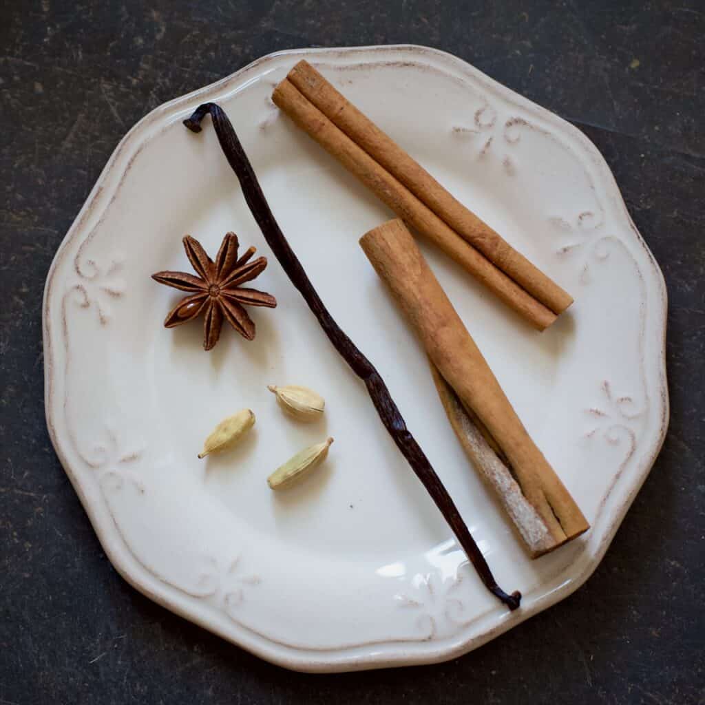 Small white plate with 2 sticks of cinnamon, 1 vanilla pod, 1 star anise and three green cardamom pods