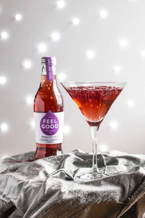 A cocktail glass of Pomagranate Popper Cocktail beside a bottle of Feel Good Cranberry and Pomagranate juice