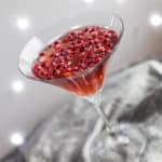 cocktail glass with pomegranate popper cocktail and pomegranate seeds against a sparkly grey background